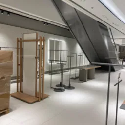 Wnętrze sklepu odzieżowego w trakcie aranżacji: metalowe stojaki na ubrania z okrągłymi podstawami, drewniany regał z jasną płytą, eskalator w tle, minimalistyczny design.