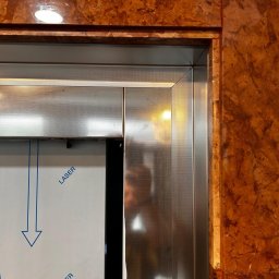 DeLift Łukasz Czechowski - Fragment metalowej konstrukcji z perforacją, zabezpieczonej folią z nadrukiem 'LASER' i strzałką, sąsiadującej z okładziną imitującą marmur.