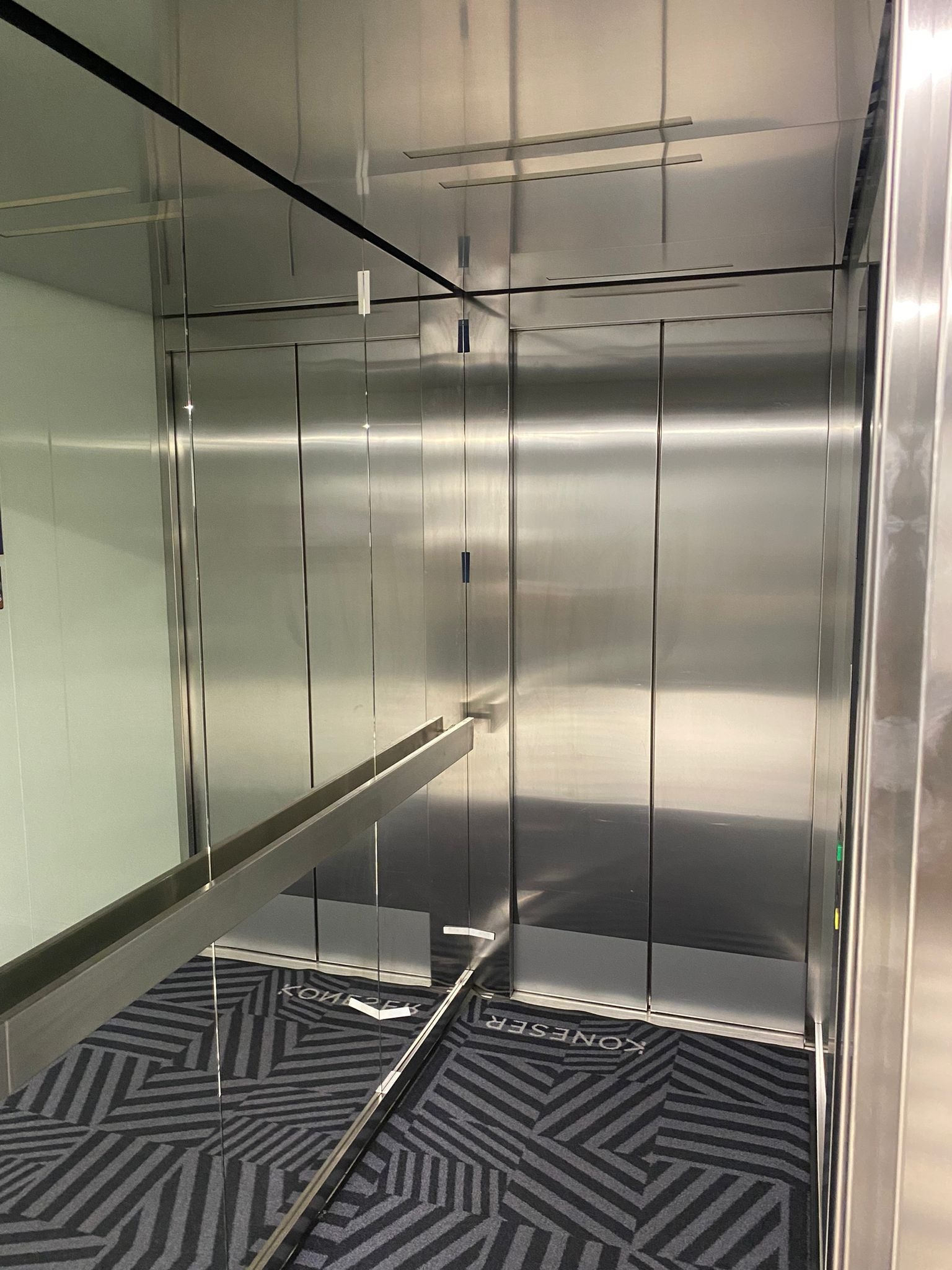 Wnętrze windy z nierdzewnej stali z poręczą i wzorzystą podłogą, widoczne odbicie w lustrzanych ścianach.