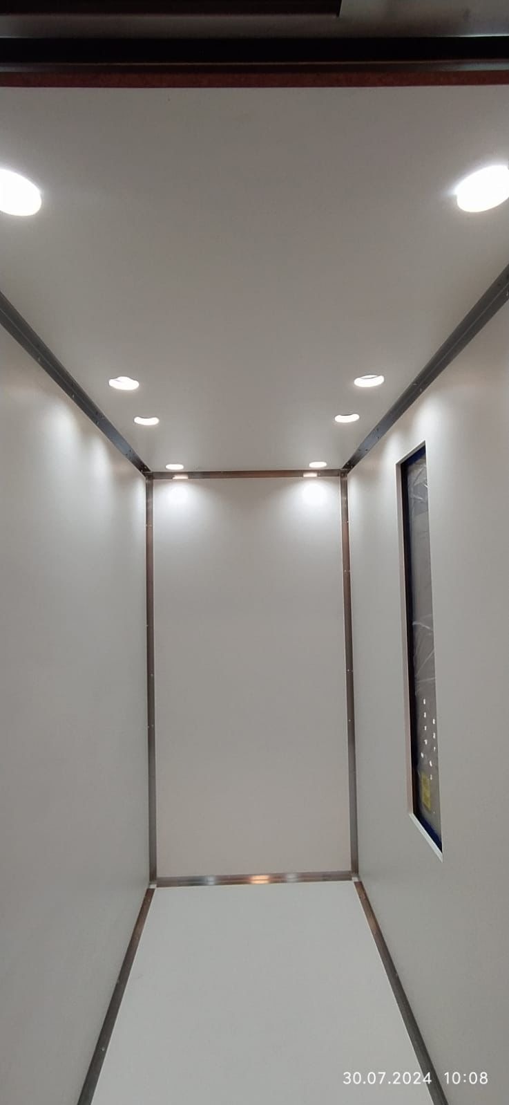 Wnętrze kabiny windy z białymi ścianami i sufitami, oświetlone punktowymi lampami LED, z metalowymi ramami wykończeniowymi i panelem sterowania po prawej stronie.