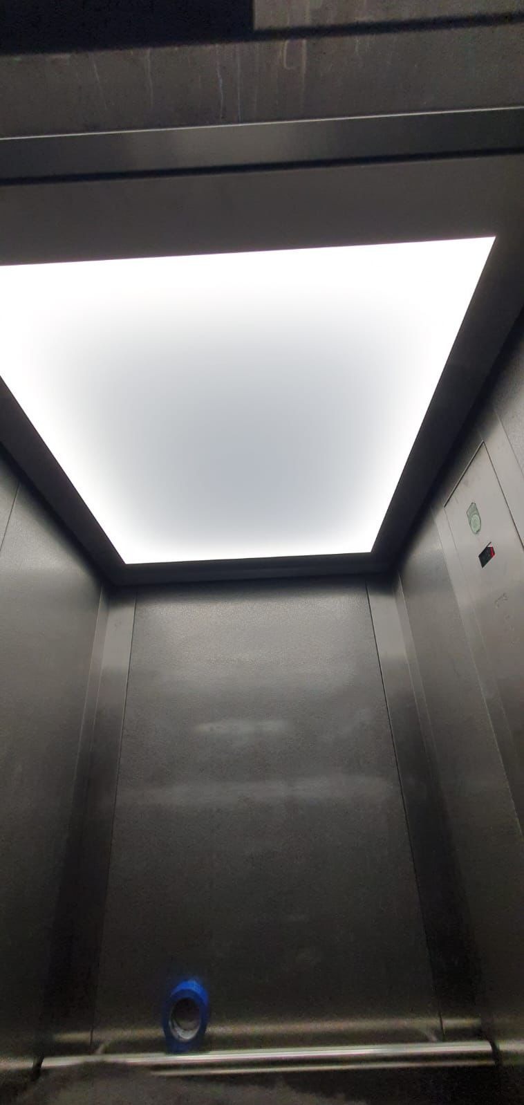 Wnętrze metalowej kabiny windy z oświetleniem sufitowym, widok z dołu, z widoczną taśmą malarską na podłodze.