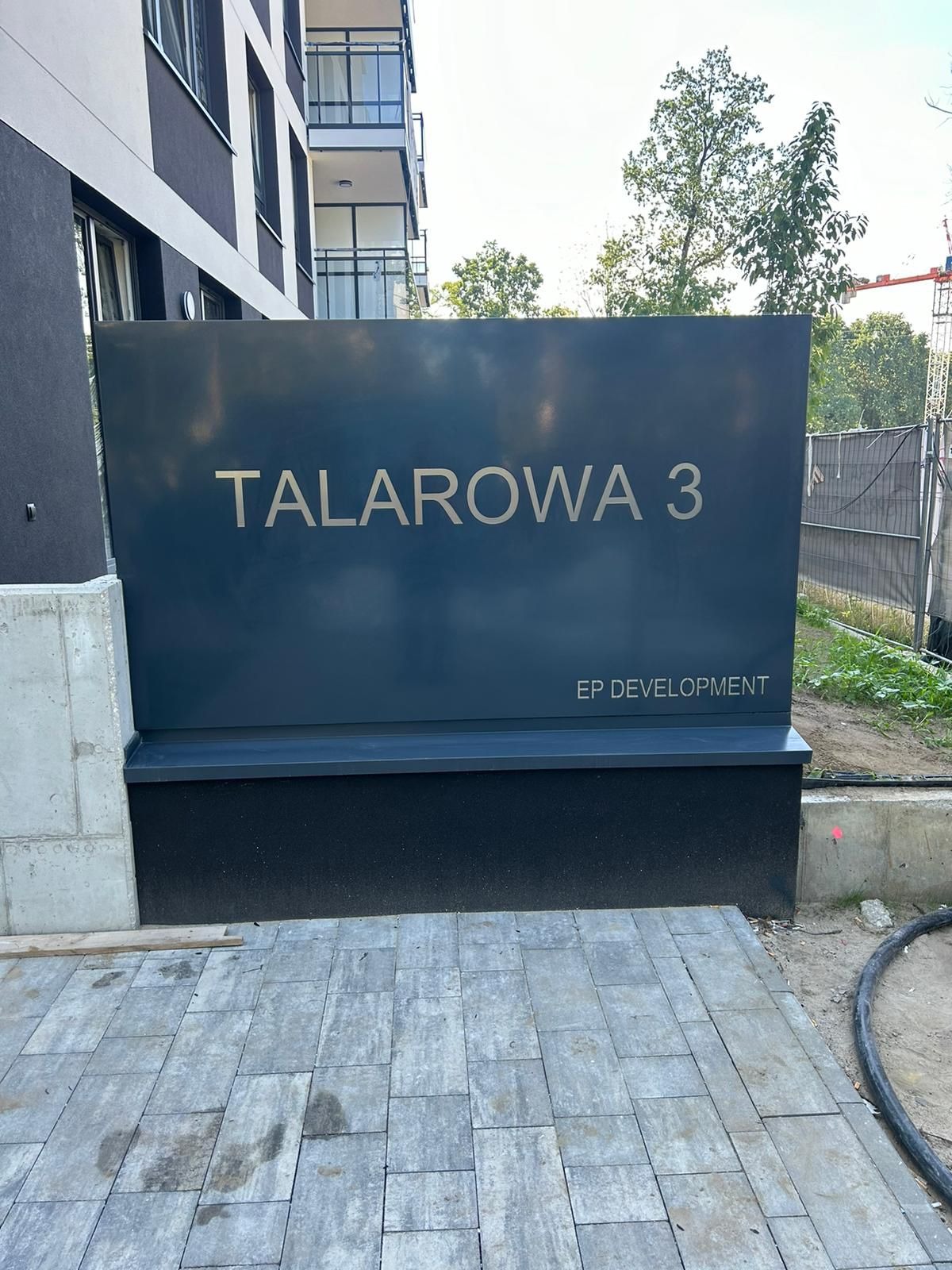 Tablica informacyjna z adresem 'Talarowa 3' i logo 'EP Development' na tle budynku mieszkalnego w trakcie budowy.