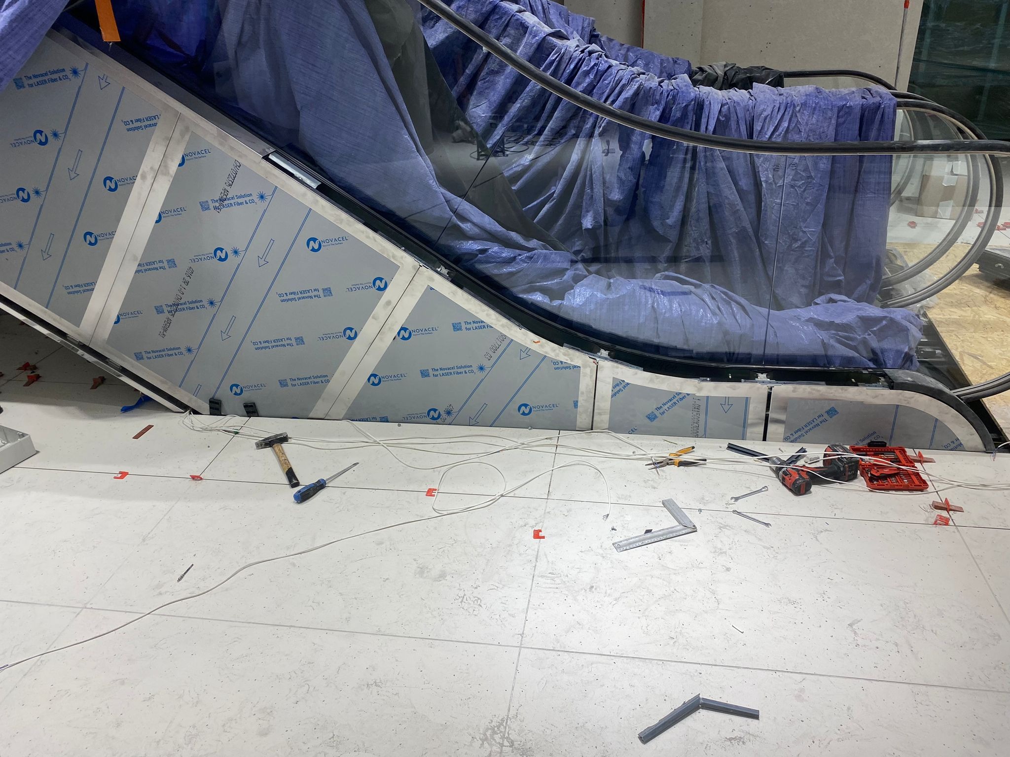 Montaż schodów ruchomych: widoczne narzędzia, elementy konstrukcyjne zabezpieczone folią ochronną Novacel, częściowo ułożona podłoga z płytek.
