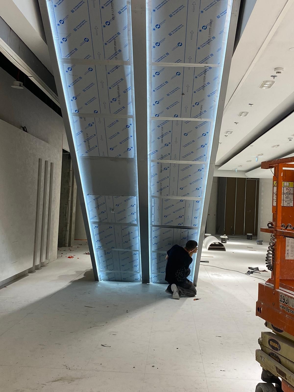 Montaż podświetlanych konstrukcji stalowych w nowoczesnym wnętrzu; pracownik na kolanach przy kolumnie, widoczny podnośnik koszowy.