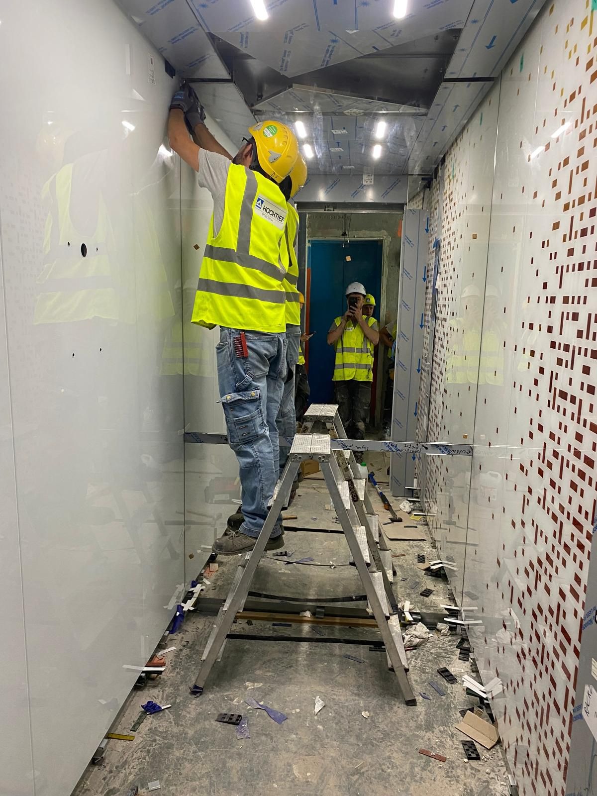 Pracownik w żółtym kasku i kamizelce odblaskowej montuje panele wewnątrz budynku, stojąc na aluminiowej drabinie, widoczne ślady po pracy na podłodze, w tle inny pracownik robi zdjęcie telefonem.