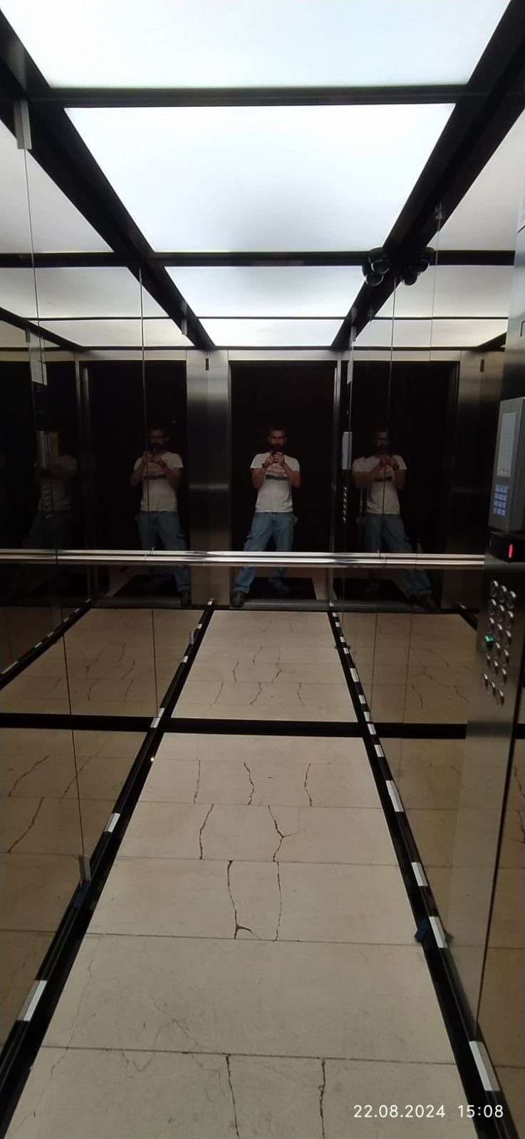 Wnętrze windy z lustrzanymi ścianami, widoczny mężczyzna robiący zdjęcie telefonem, marmurowa podłoga w neutralnych barwach, oświetlenie sufitowe w czarnej ramie.