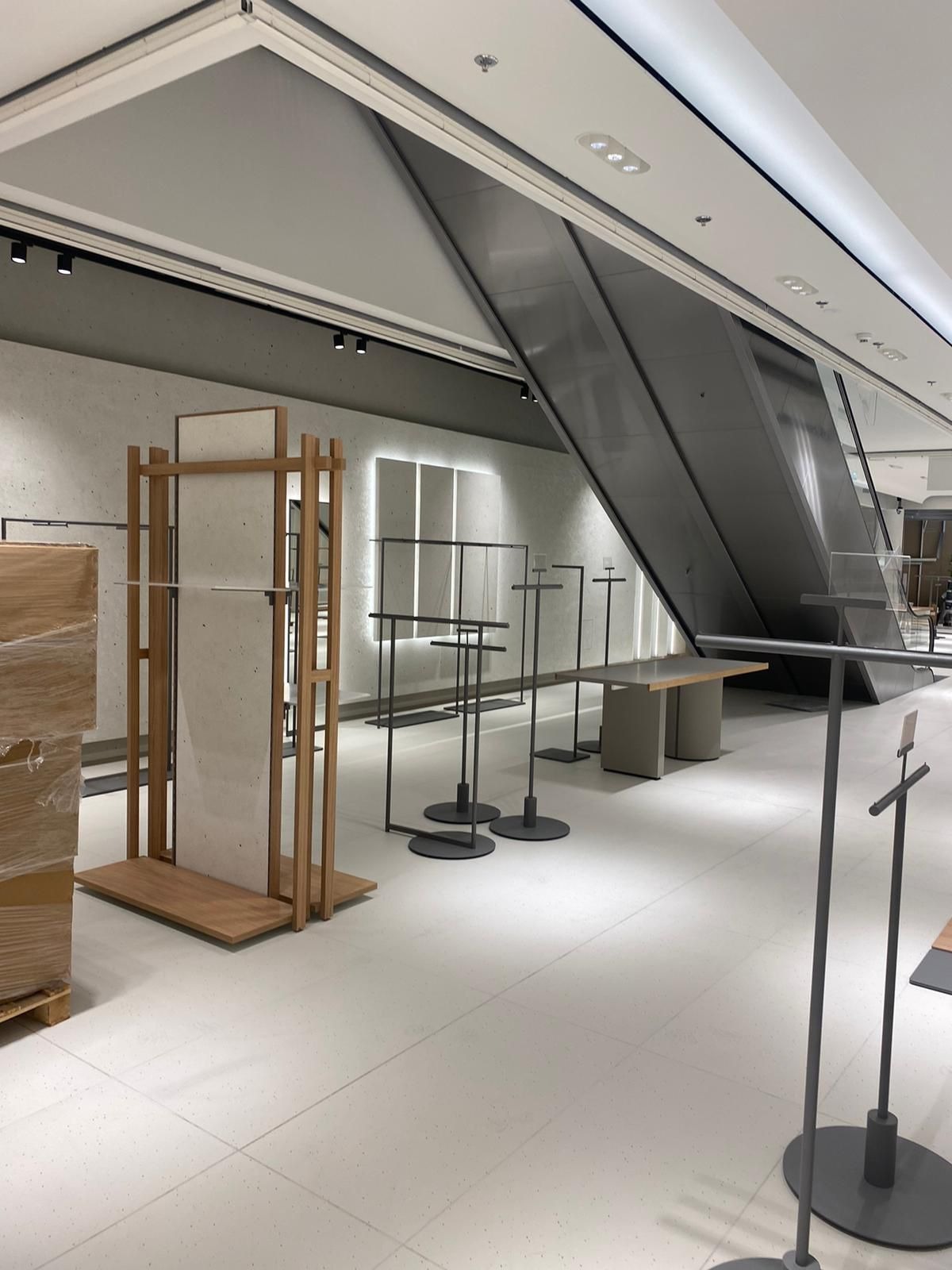 Wnętrze sklepu odzieżowego w trakcie aranżacji: metalowe stojaki na ubrania z okrągłymi podstawami, drewniany regał z jasną płytą, eskalator w tle, minimalistyczny design.