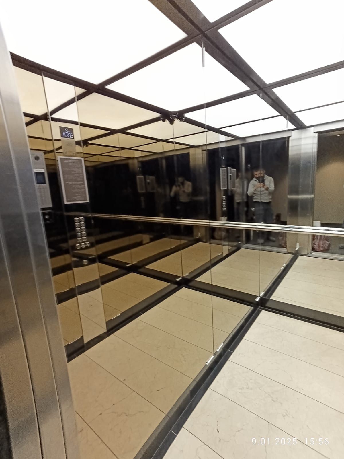 Wnętrze windy z lustrzanymi ścianami, widoczny panel sterowania i odbicie osoby robiącej zdjęcie, jasne oświetlenie sufitowe.