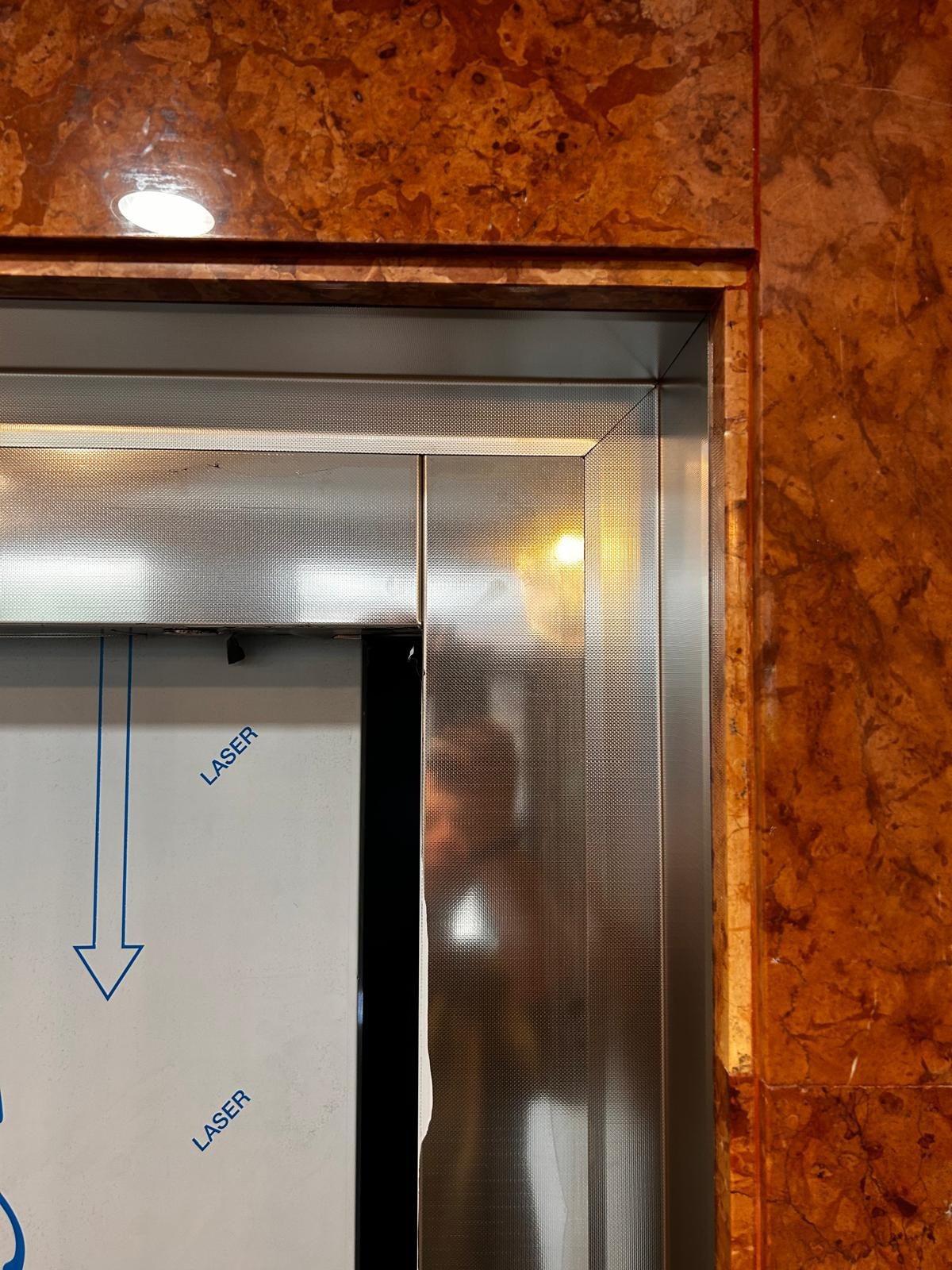 Fragment metalowej konstrukcji z perforacją, zabezpieczonej folią z nadrukiem 'LASER' i strzałką, sąsiadującej z okładziną imitującą marmur.