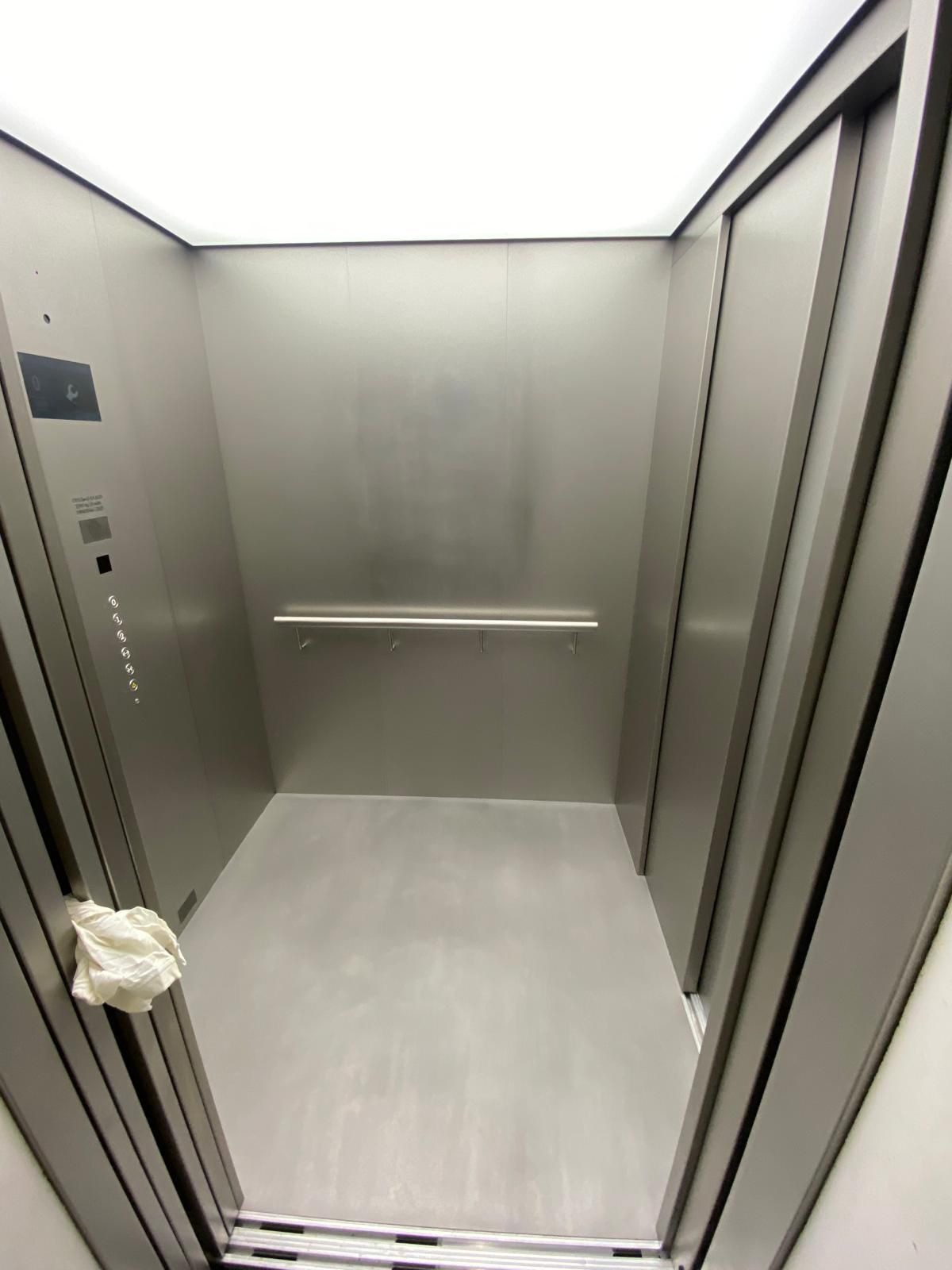Wnętrze windy z metalowymi ścianami, oświetleniem LED i panelem sterowania po lewej stronie, widok z dołu.