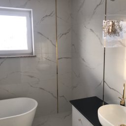 Jacek Czarnecki - Nowoczesna łazienka z białymi płytkami imitującymi marmur, złotymi listwami dekoracyjnymi, wolnostojącą wanną i umywalką nablatową z czarnym blatem.