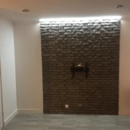 Ściana z cegły dekoracyjnej z zamontowanym uchwytem do telewizora, oświetlona listwą LED, na podłodze panele imitujące drewno.