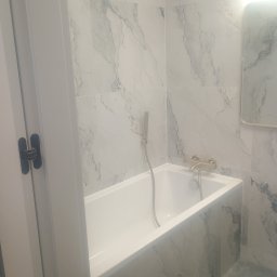 Jacek Czarnecki - Biała wanna w łazience wyłożonej marmurowymi płytkami, z widoczną armaturą w kolorze złota i fragmentem lustra. Minimalistyczny design.