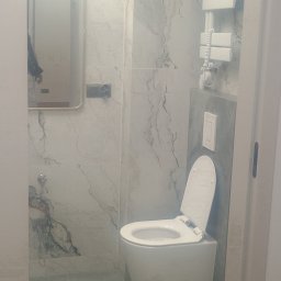 Jacek Czarnecki - Nowoczesna łazienka z marmurowymi płytkami, podwieszaną toaletą i grzejnikiem drabinkowym. Lustro z podświetleniem odbija fragment osoby.