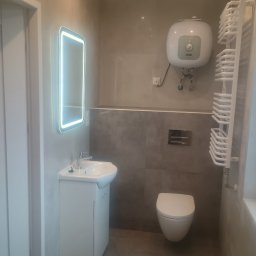 Jacek Czarnecki - Wykończona łazienka w Duczkach z podświetlanym lustrem, umywalką z szafką, toaletą i grzejnikiem drabinkowym. Szare płytki na ścianach i podłodze.