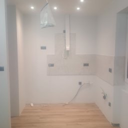 Jacek Czarnecki - Wnętrze kuchni w trakcie remontu: ułożone panele podłogowe, częściowo położone płytki na ścianie, widoczne gniazdka elektryczne i instalacja wodna.