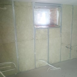 Jacek Czarnecki - Ściana z izolacją z wełny mineralnej, zamontowana na metalowym stelażu, widoczne przewody elektryczne i okno z ceglanym obramowaniem. Etap prac wykończeniowych.