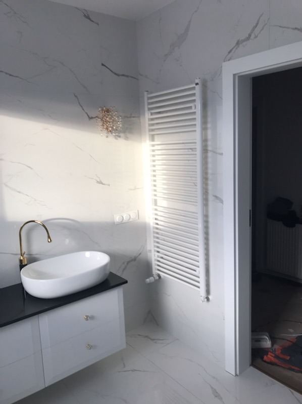 Nowoczesna łazienka z umywalką nablatową, złotą baterią i białym grzejnikiem drabinkowym na tle marmurowych płytek.