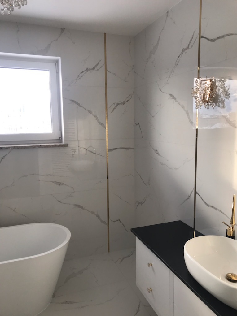 Nowoczesna łazienka z białymi płytkami imitującymi marmur, złotymi listwami dekoracyjnymi, wolnostojącą wanną i umywalką nablatową z czarnym blatem.