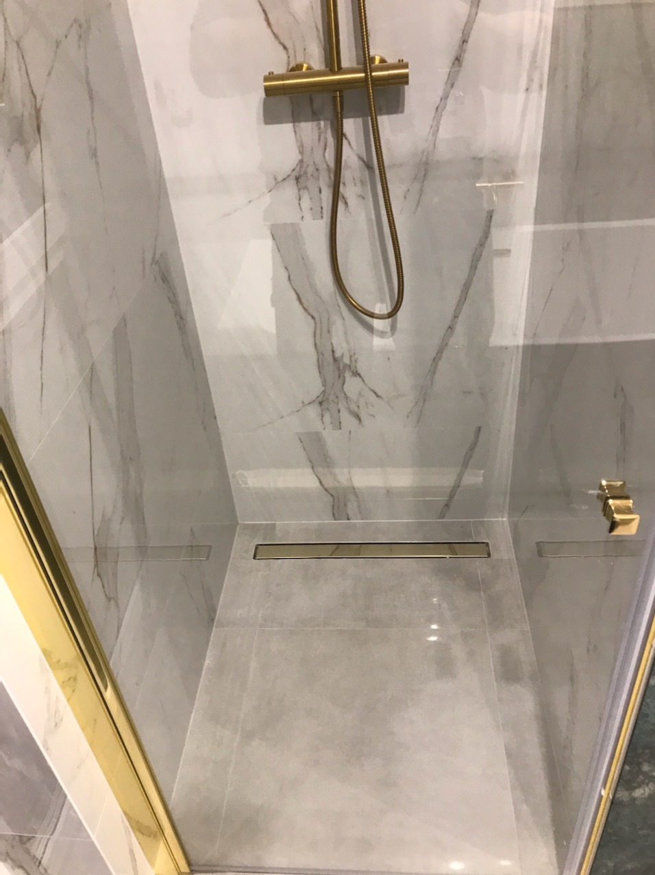 Nowoczesna kabina prysznicowa z marmurowymi płytkami, złotą armaturą i odpływem liniowym w podłodze.