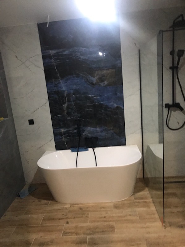 Nowoczesna łazienka z wolnostojącą białą wanną, ciemną płytą imitującą marmur na ścianie i kabiną prysznicową ze szkła.