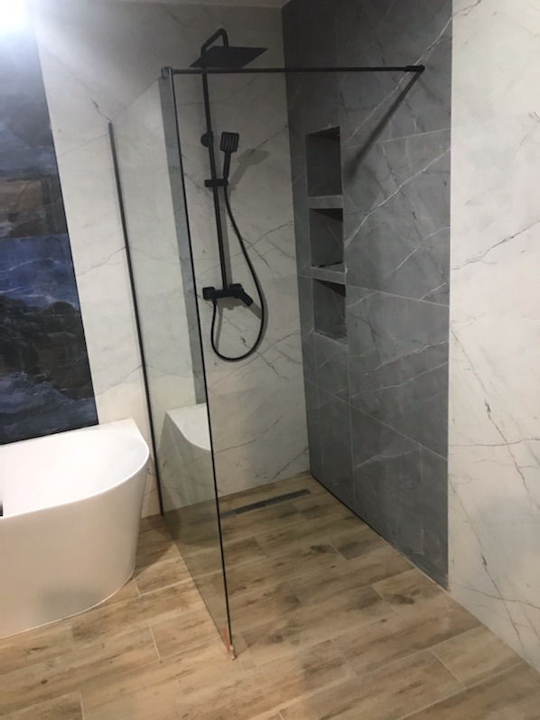 Nowoczesna łazienka z kabiną prysznicową typu walk-in, czarną armaturą, marmurowymi płytkami i drewnopodobną podłogą. W tle widoczna wanna.