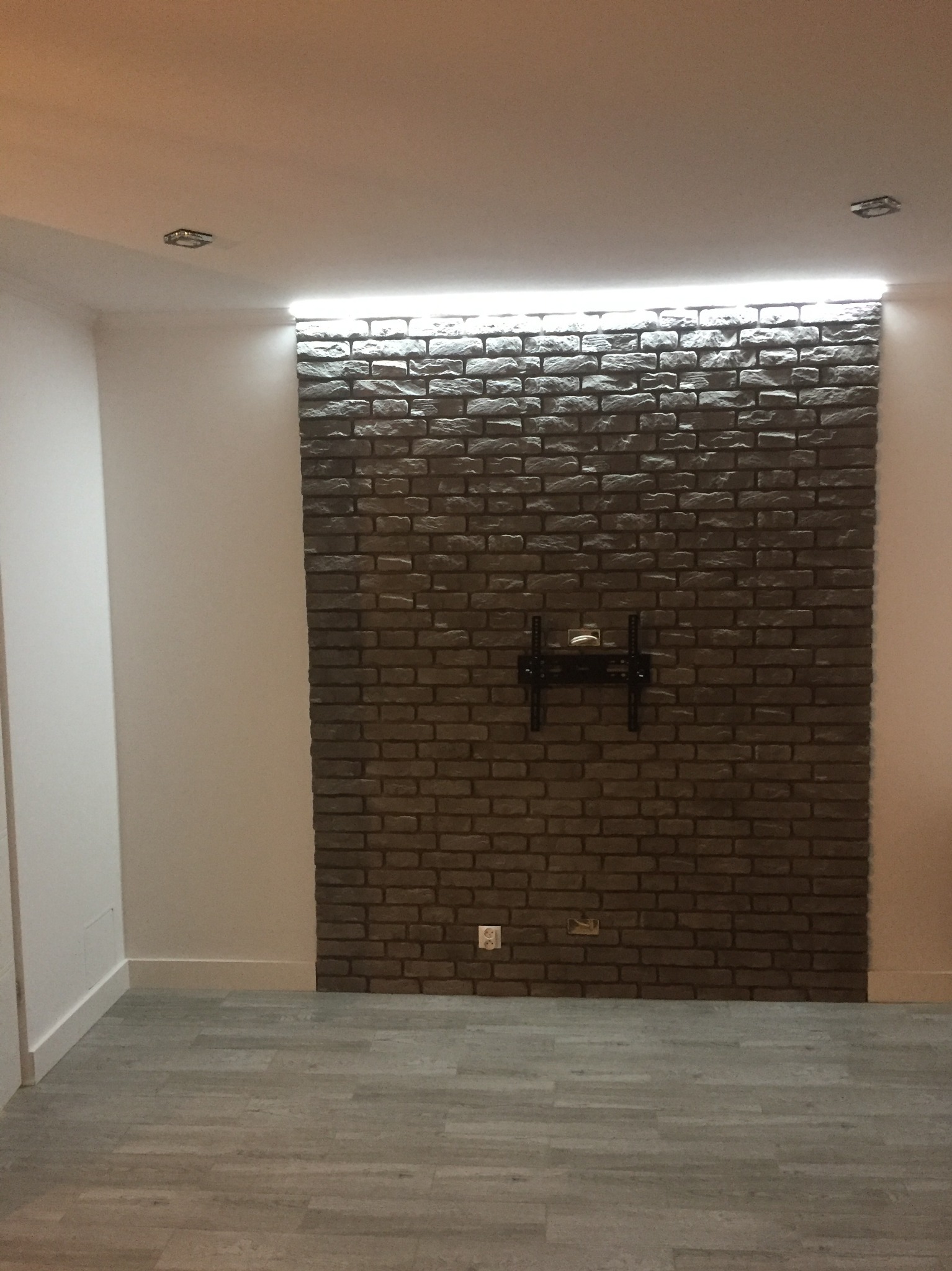 Ściana z cegły dekoracyjnej z zamontowanym uchwytem do telewizora, oświetlona listwą LED, na podłodze panele imitujące drewno.