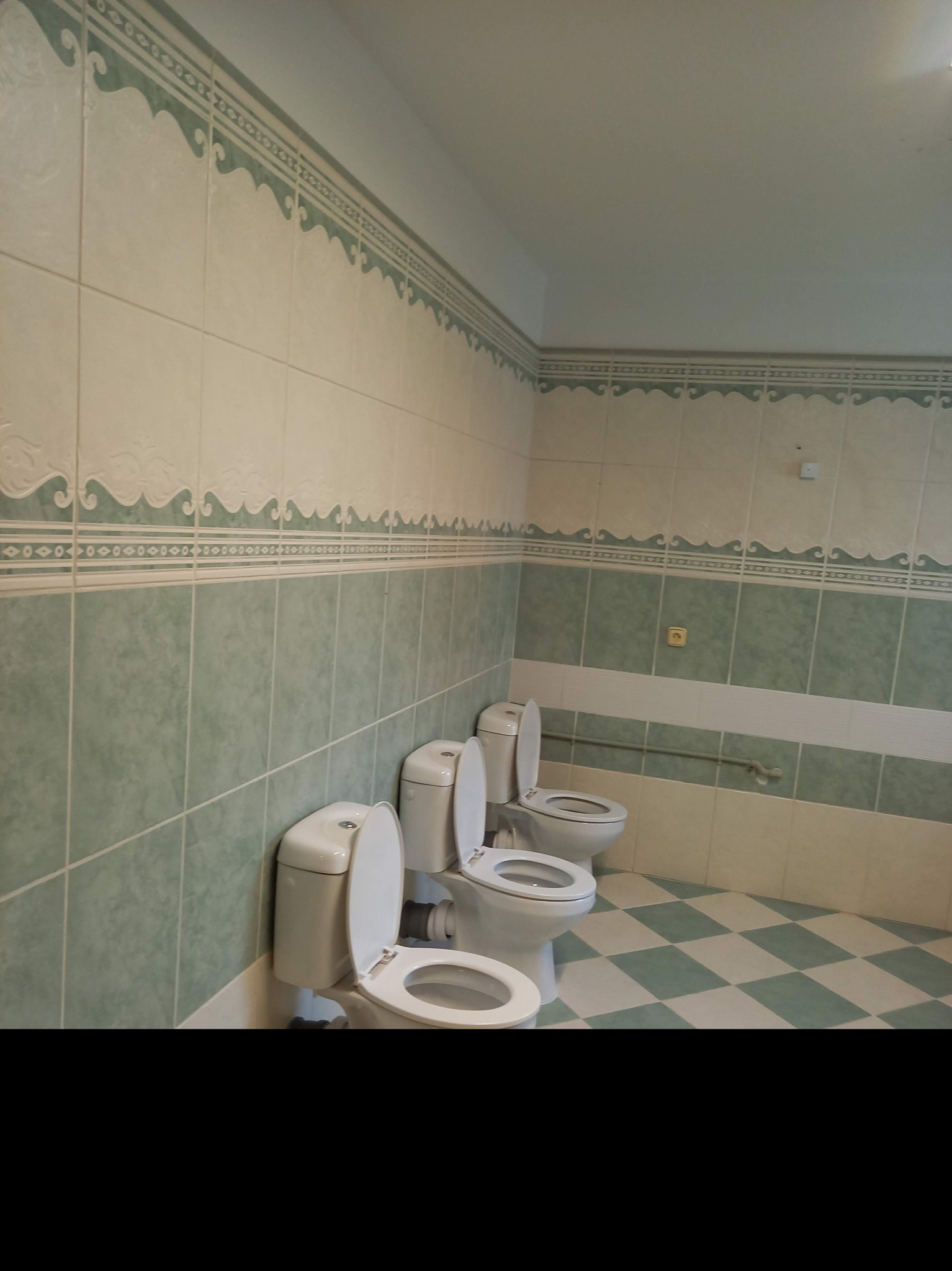 Trzy białe toalety ustawione jedna za drugą w wykafelkowanej łazience z zielono-białą szachownicą na podłodze i dekoracyjnym pasem płytek na ścianach.