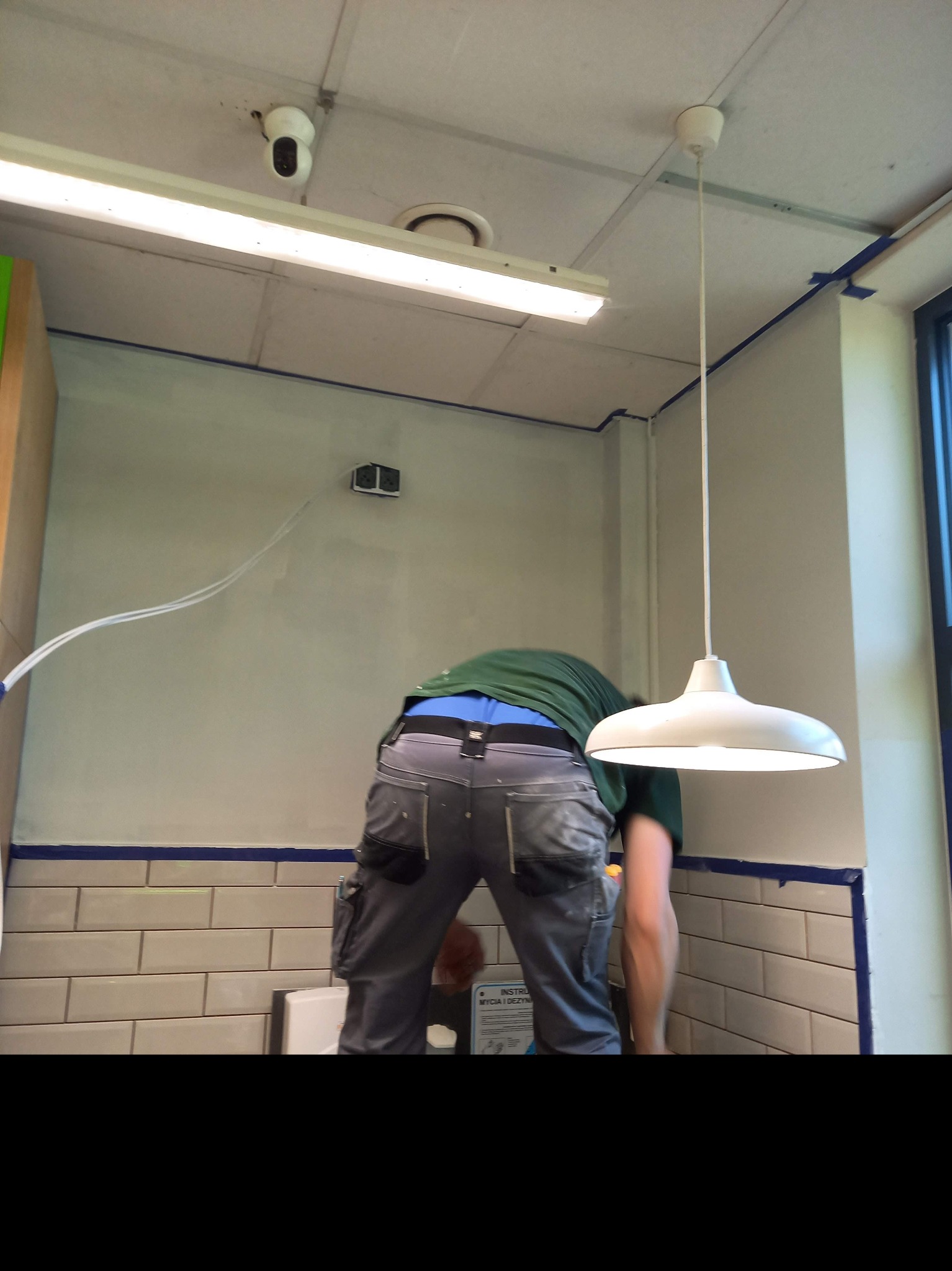 Pracownik pochylony nad ścianą w trakcie malowania, zabezpieczona niebieską taśmą malarską, widoczna biała lampa wisząca i płytki na ścianie.