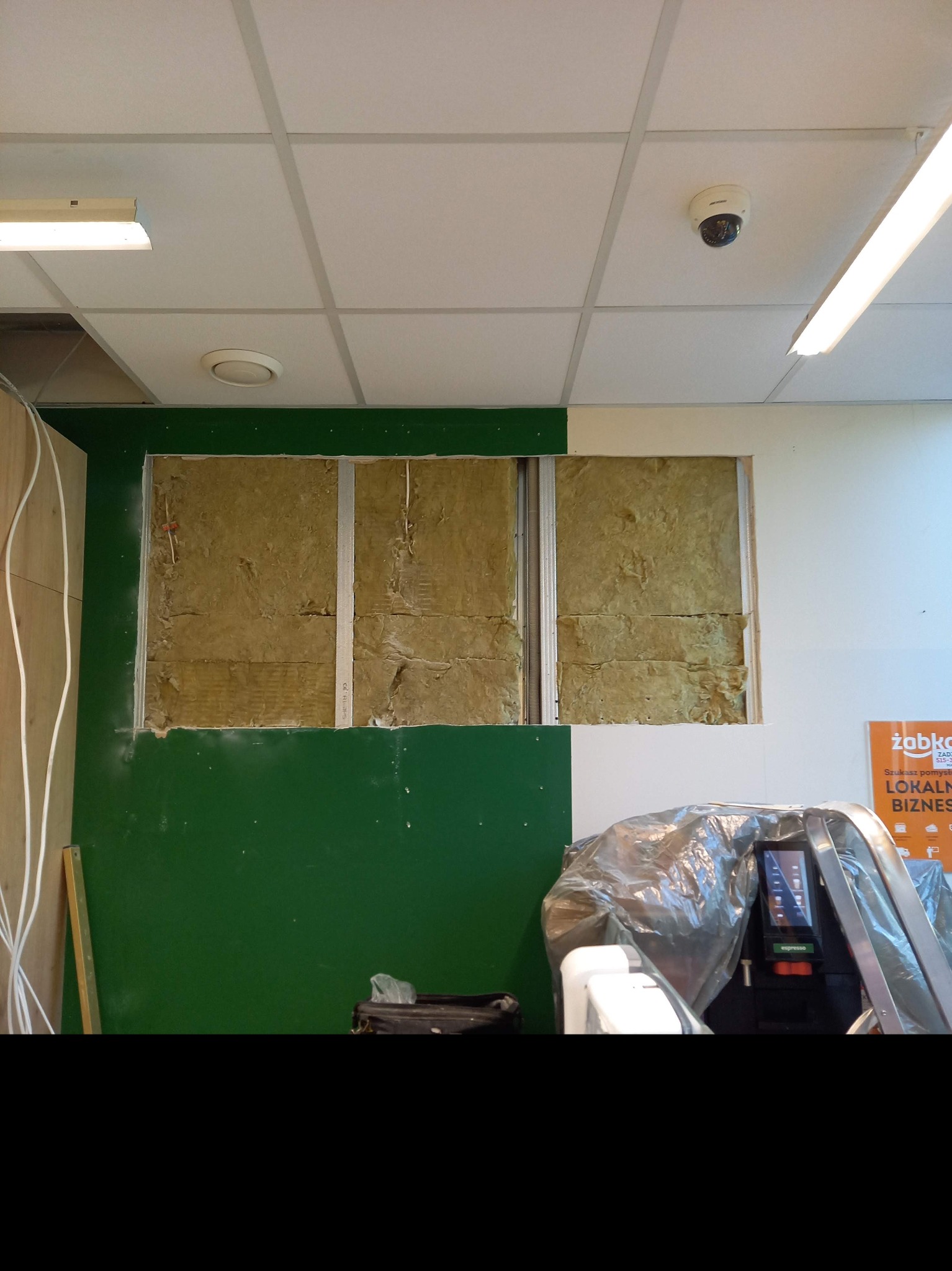 Ściana w trakcie ocieplania wełną mineralną w lokalu w Duczkach, widoczne profile metalowe, zielona farba na ścianie, sufit kasetonowy z oświetleniem i kamerą.