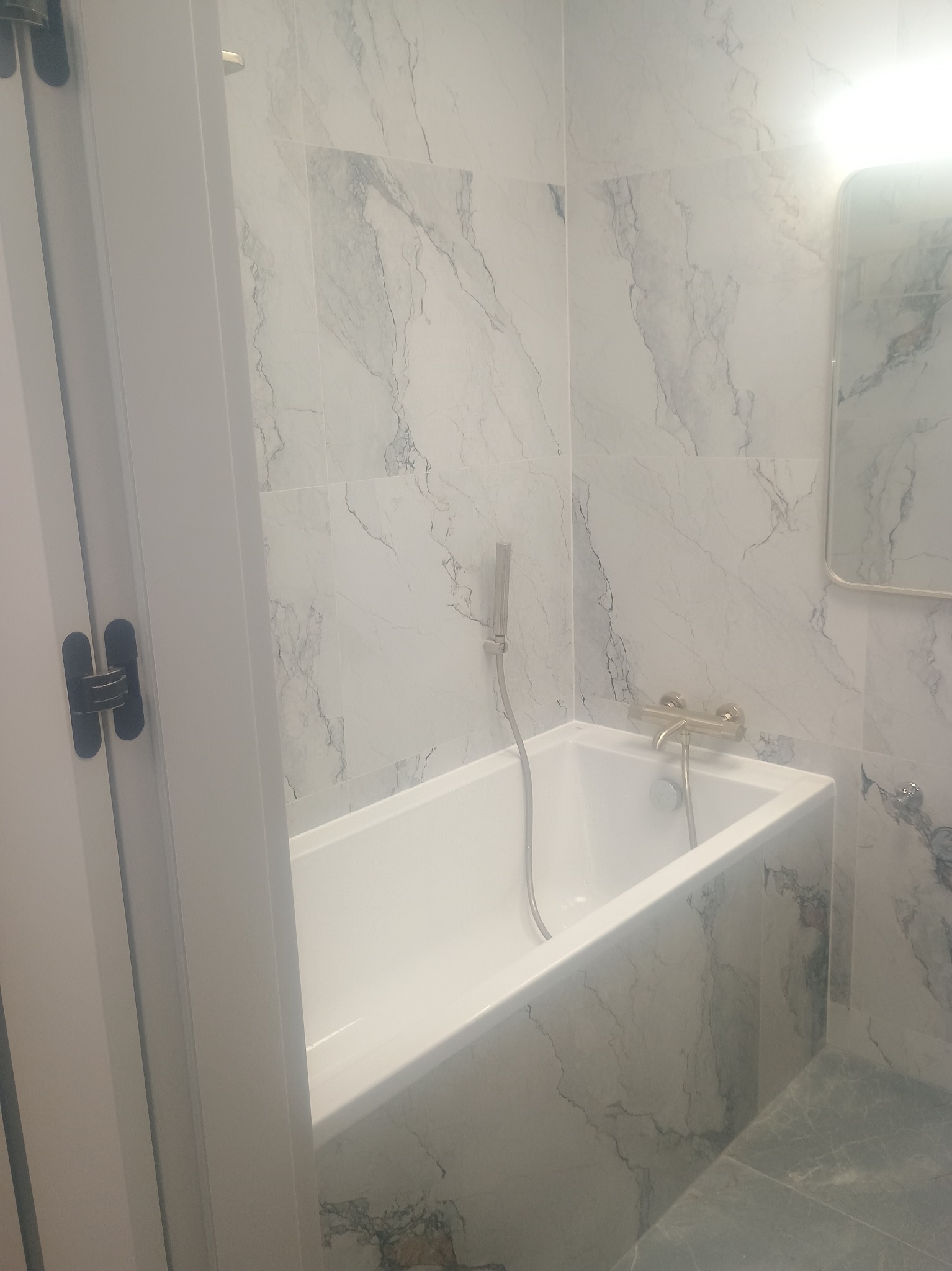 Biała wanna w łazience wyłożonej marmurowymi płytkami, z widoczną armaturą w kolorze złota i fragmentem lustra. Minimalistyczny design.