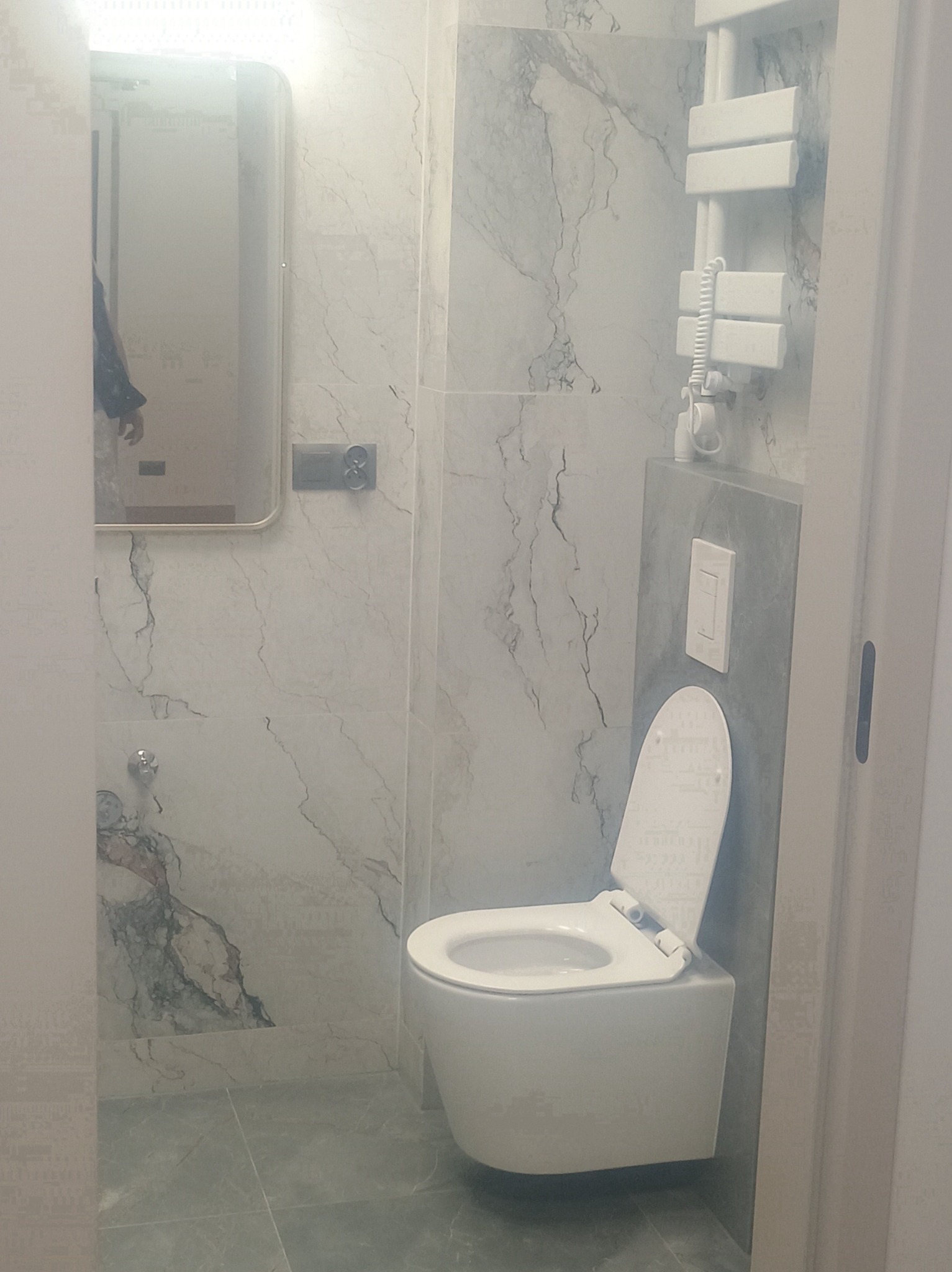 Nowoczesna łazienka z marmurowymi płytkami, podwieszaną toaletą i grzejnikiem drabinkowym. Lustro z podświetleniem odbija fragment osoby.