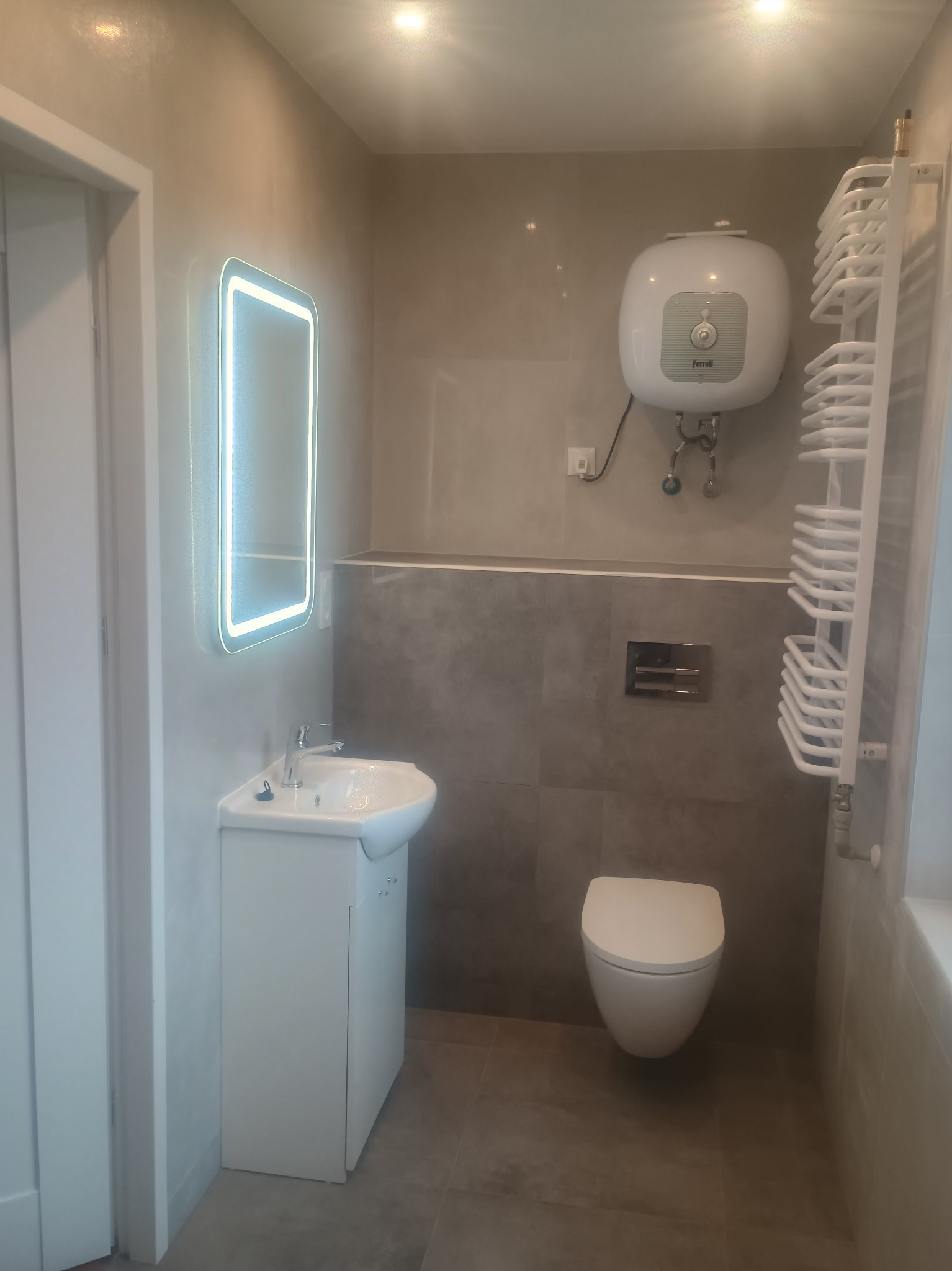 Wykończona łazienka w Duczkach z podświetlanym lustrem, umywalką z szafką, toaletą i grzejnikiem drabinkowym. Szare płytki na ścianach i podłodze.