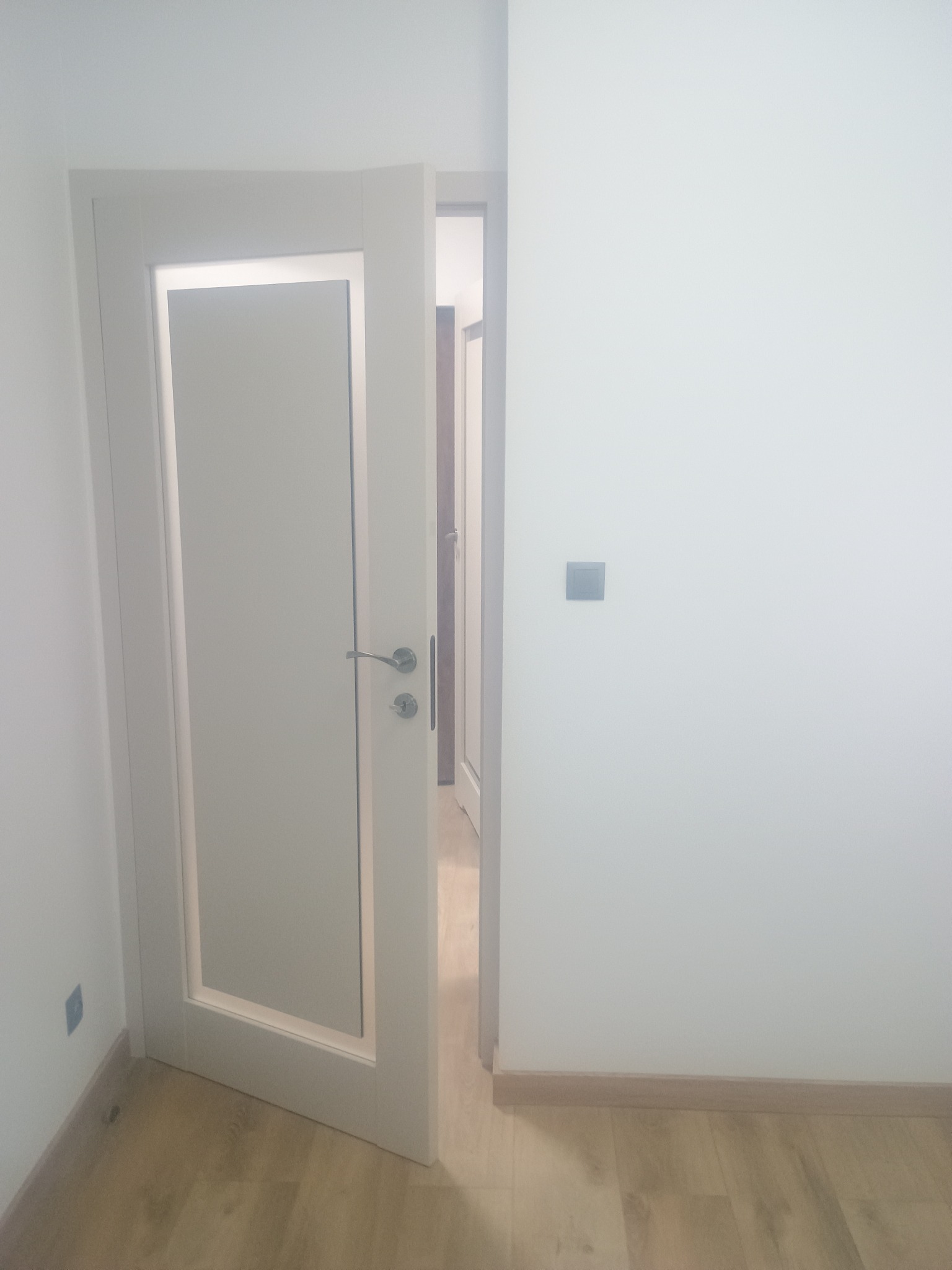 Otwarta, biała, nowoczesna stolarka drzwiowa z przeszkleniem w minimalistycznym wnętrzu. Widoczny fragment podłogi i ściany z włącznikiem.