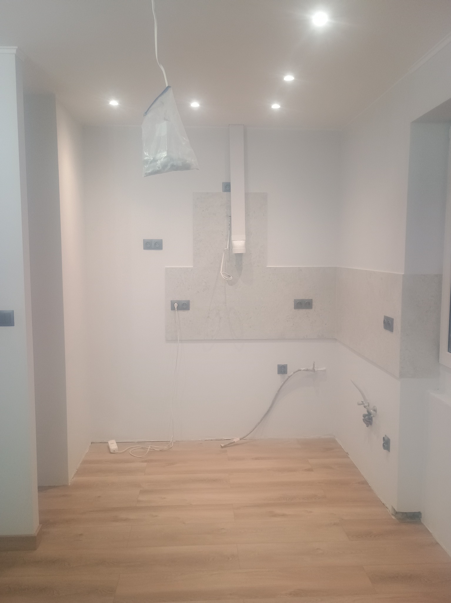 Wnętrze kuchni w trakcie remontu: ułożone panele podłogowe, częściowo położone płytki na ścianie, widoczne gniazdka elektryczne i instalacja wodna.