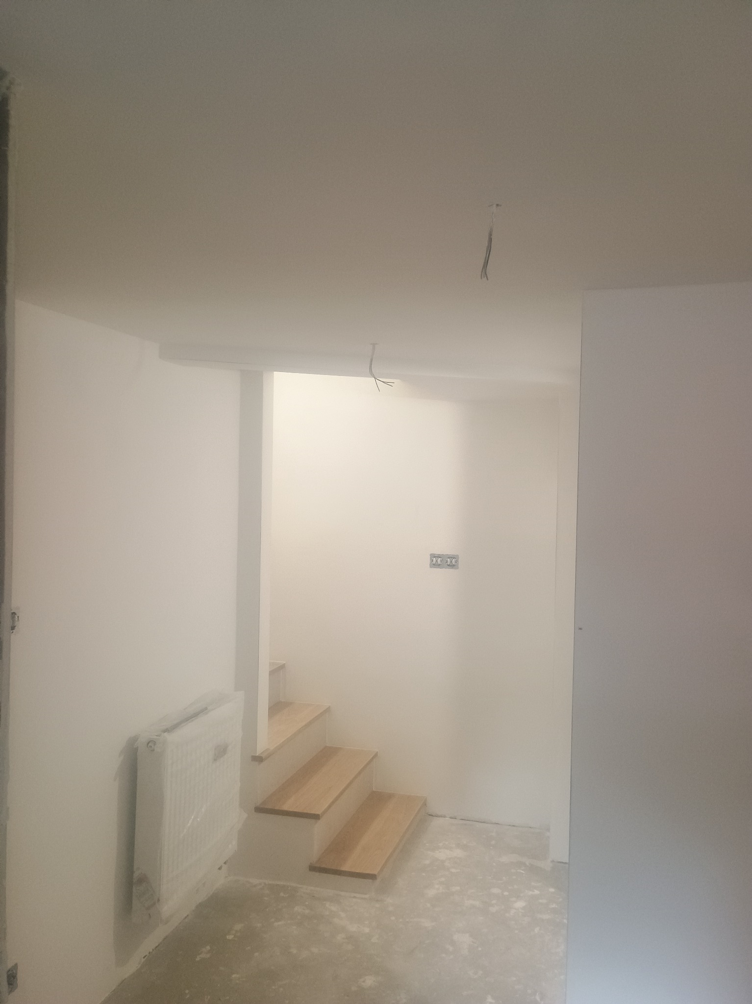 Wnętrze w trakcie wykańczania w Duczkach: świeżo pomalowane ściany na biało, drewniane schody, grzejnik zabezpieczony folią, surowa podłoga.