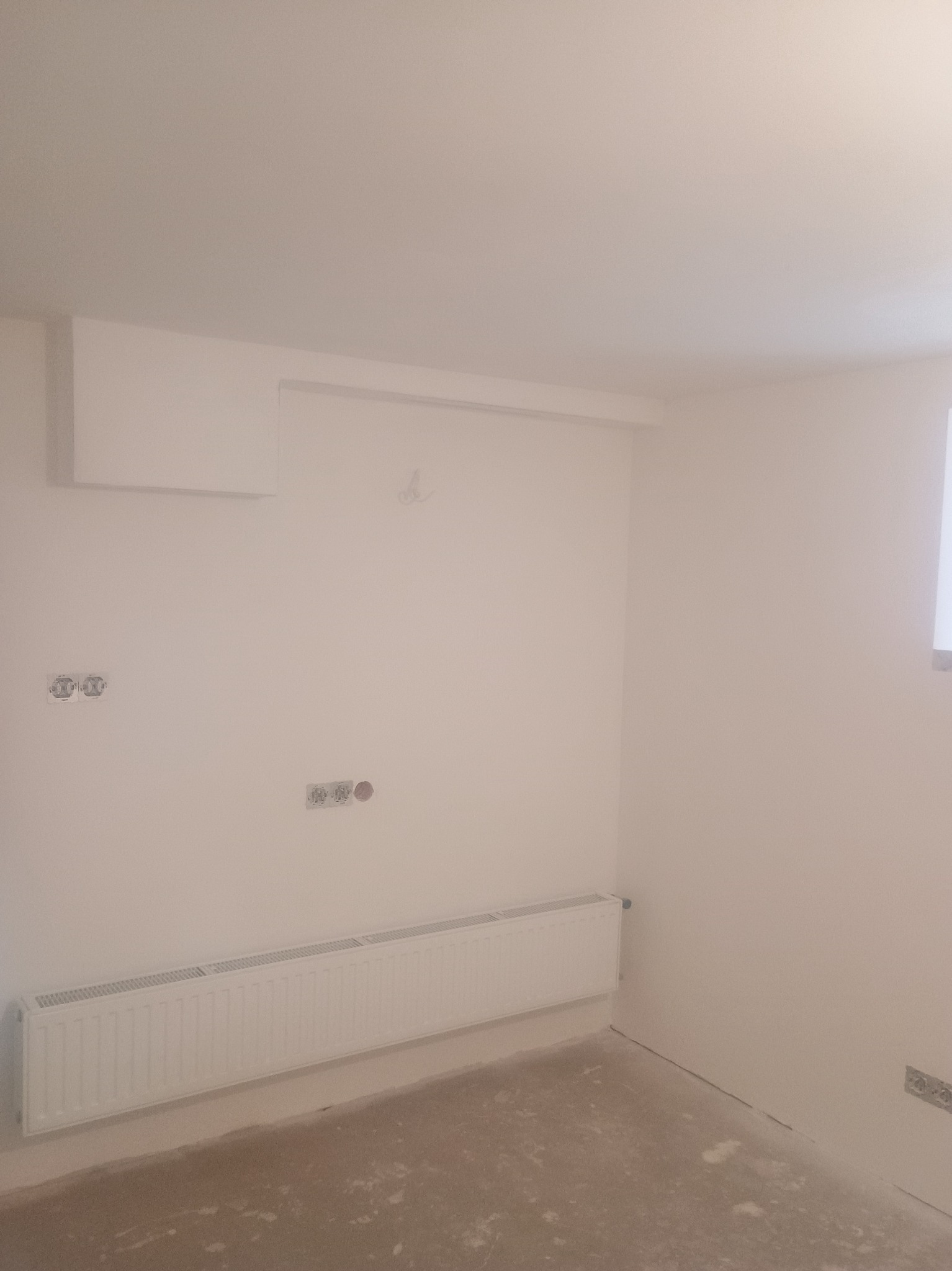 Pomalowane na biało wnętrze pokoju z grzejnikiem, gniazdkami elektrycznymi i przygotowanym podłożem pod podłogę. Widoczne elementy instalacji.