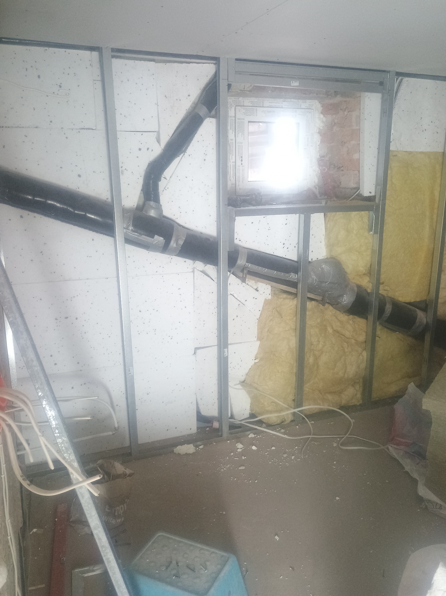 Konstrukcja szkieletowa z profili stalowych, częściowo wypełniona izolacją i instalacją wentylacyjną. Widoczne przewody i okno w surowym stanie.