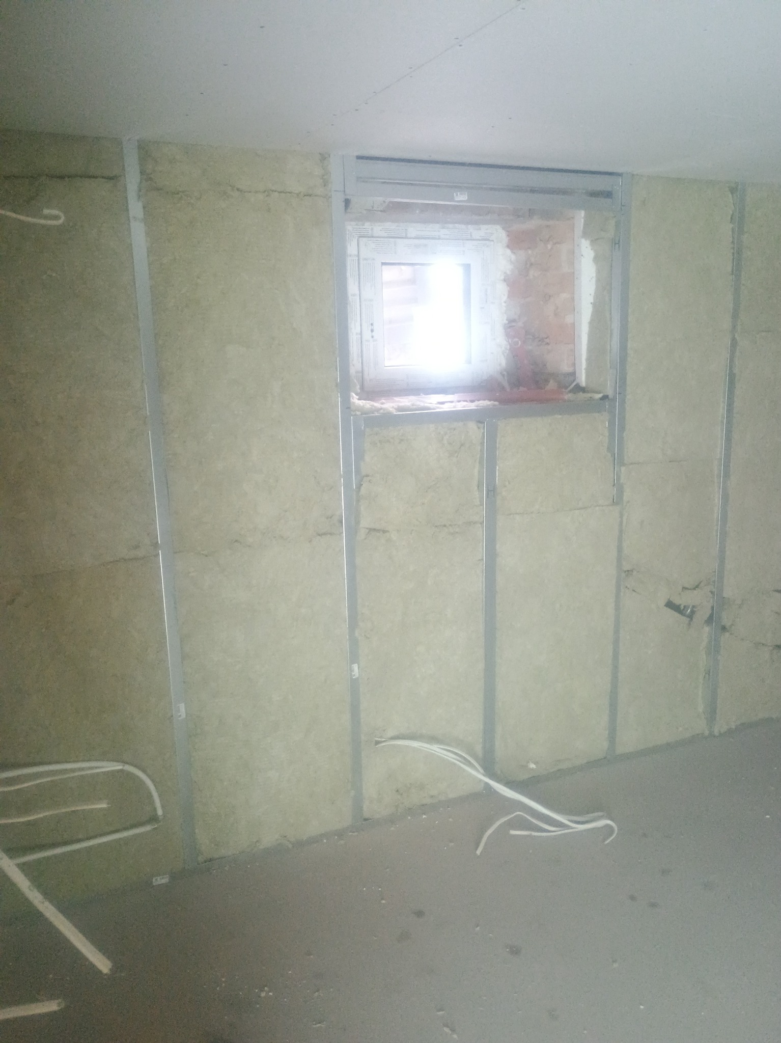 Ściana z izolacją z wełny mineralnej, zamontowana na metalowym stelażu, widoczne przewody elektryczne i okno z ceglanym obramowaniem. Etap prac wykończeniowych.
