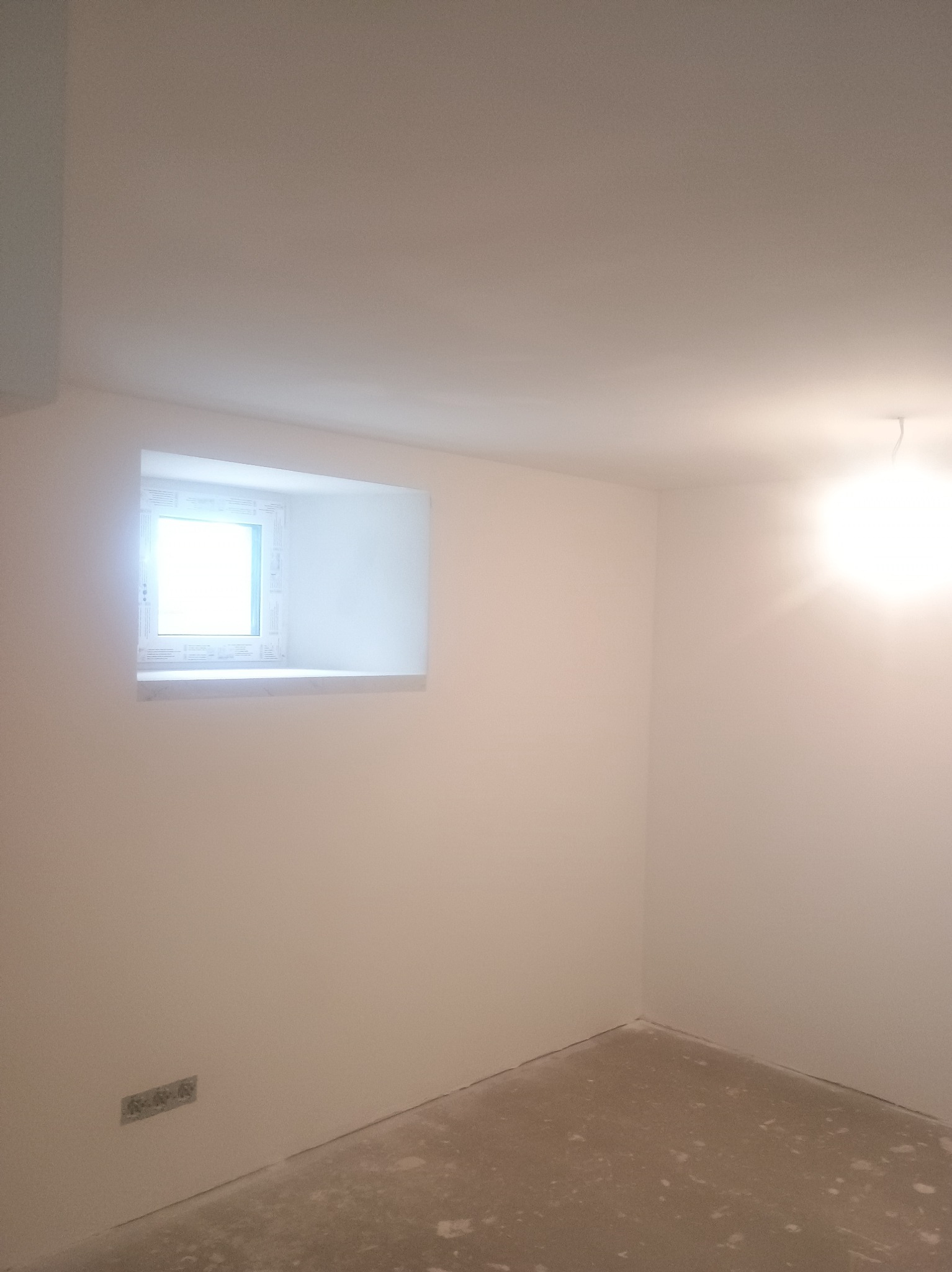 Pomalowane na biało wnętrze pokoju z oknem w ścianie. Na podłodze widoczne ślady po pracach wykończeniowych. Świeżo wykończone ściany i sufit.