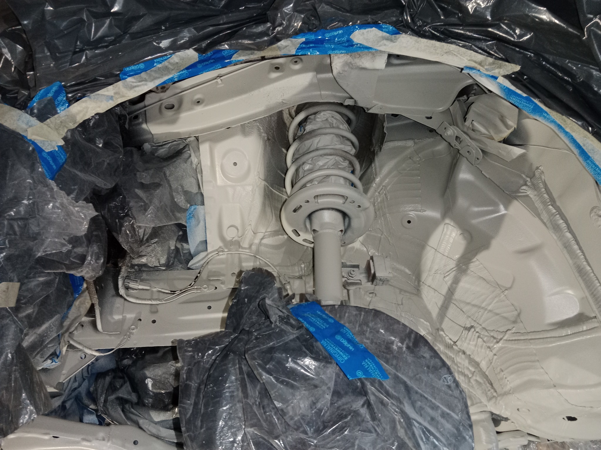 Podwozie samochodu po nałożeniu białej powłoki ochronnej, widoczny amortyzator, zabezpieczone folią i taśmą elementy karoserii.