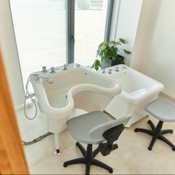 Centrum Rehabilitacji i Opieki Leśna Ostoja - Profesjonalne wanny do hydromasażu z chromowanymi bateriami i dwoma krzesłami na kółkach w jasnym gabinecie z oknem i rośliną doniczkową.