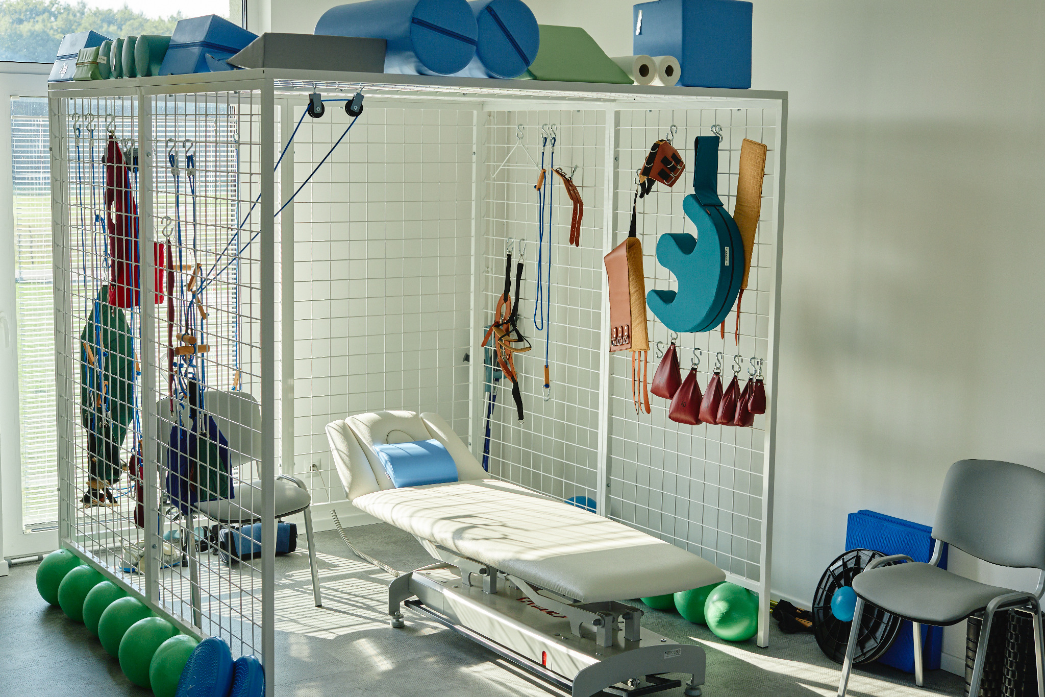 Jasny pokój z metalową klatką rehabilitacyjną, wewnątrz biały stół do ćwiczeń z niebieską poduszką, na ściankach klatki zawieszone różnorodne przybory rehabilitacyjne, na górze i na podłodze...