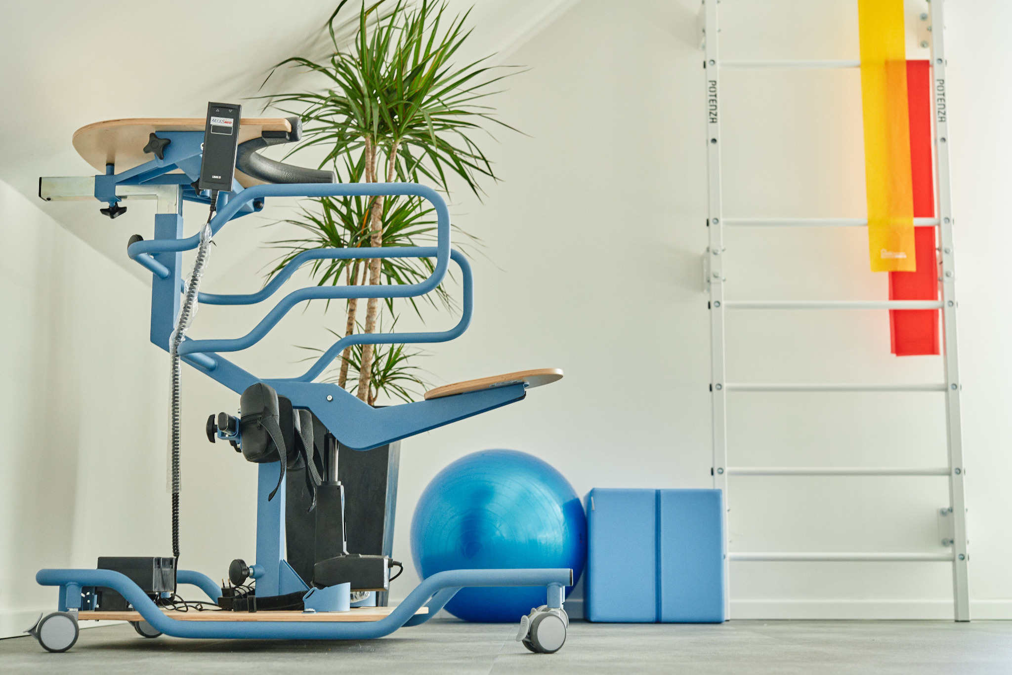 Niebieski pionizator statyczny z regulowanym podparciem, piłka rehabilitacyjna, drabinka gimnastyczna z gumami oporowymi w jasnym wnętrzu gabinetu.