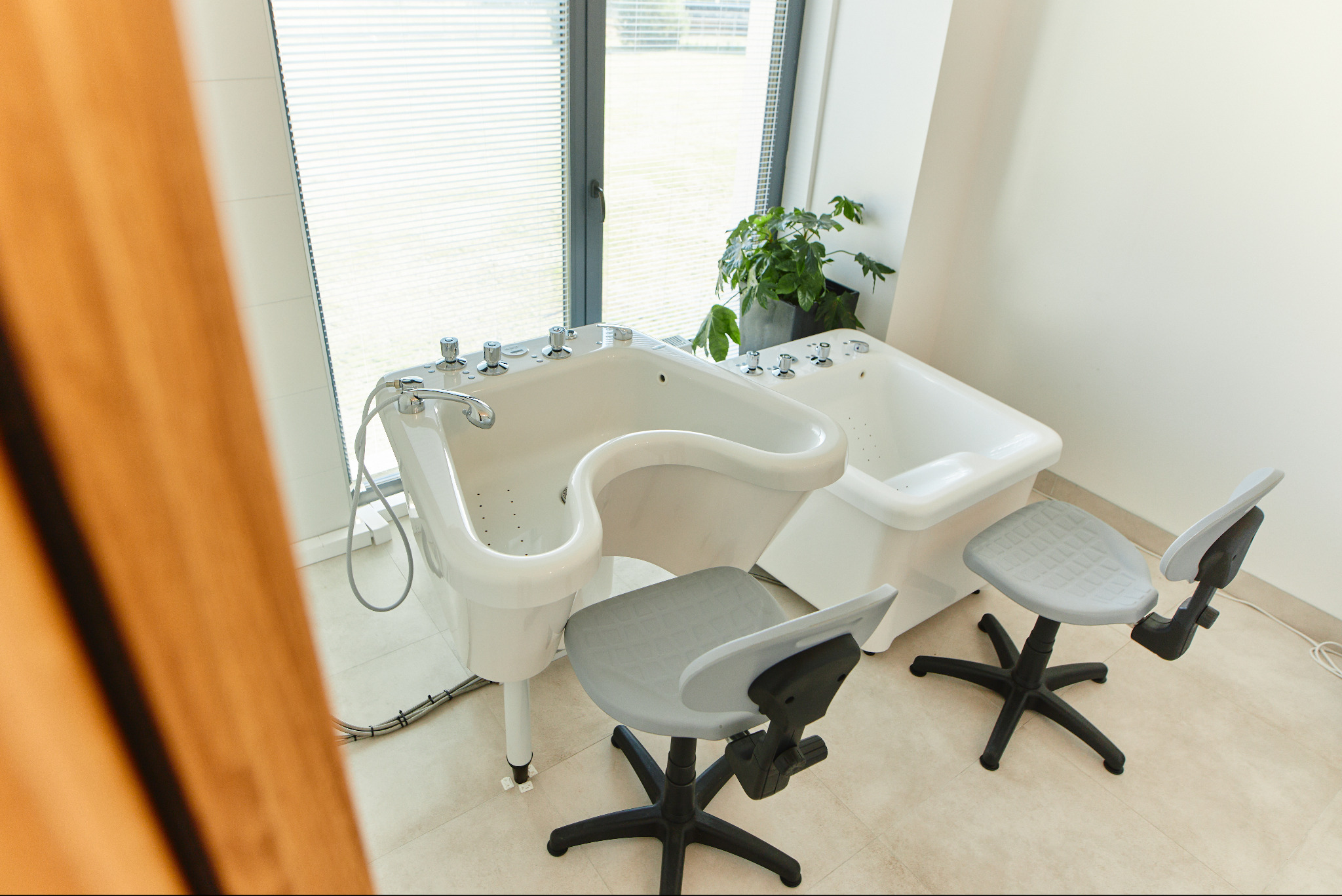 Profesjonalne wanny do hydromasażu z chromowanymi bateriami i dwoma krzesłami na kółkach w jasnym gabinecie z oknem i rośliną doniczkową.