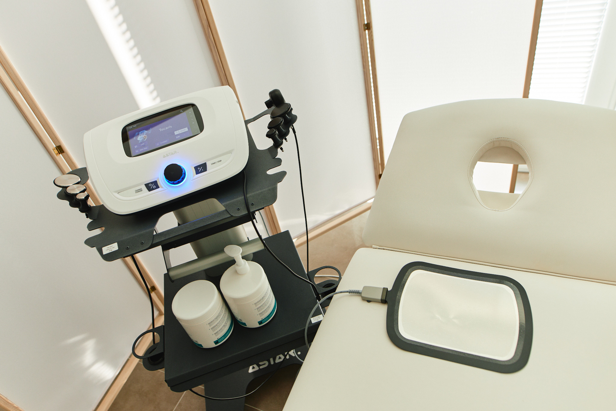 Aparat do fizykoterapii Tecaris z elektrodą na leżance, obok butelki z żelem kontaktowym i akcesoriami, w gabinecie zabiegowym.