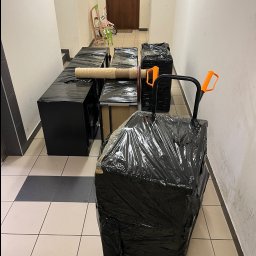 MATT Przeprowadzki Kraków, Transport - Meble i inne przedmioty zabezpieczone folią i przygotowane do transportu, ustawione w korytarzu bloku mieszkalnego, widoczny wózek transportowy z pomarańczowymi uchwytami.