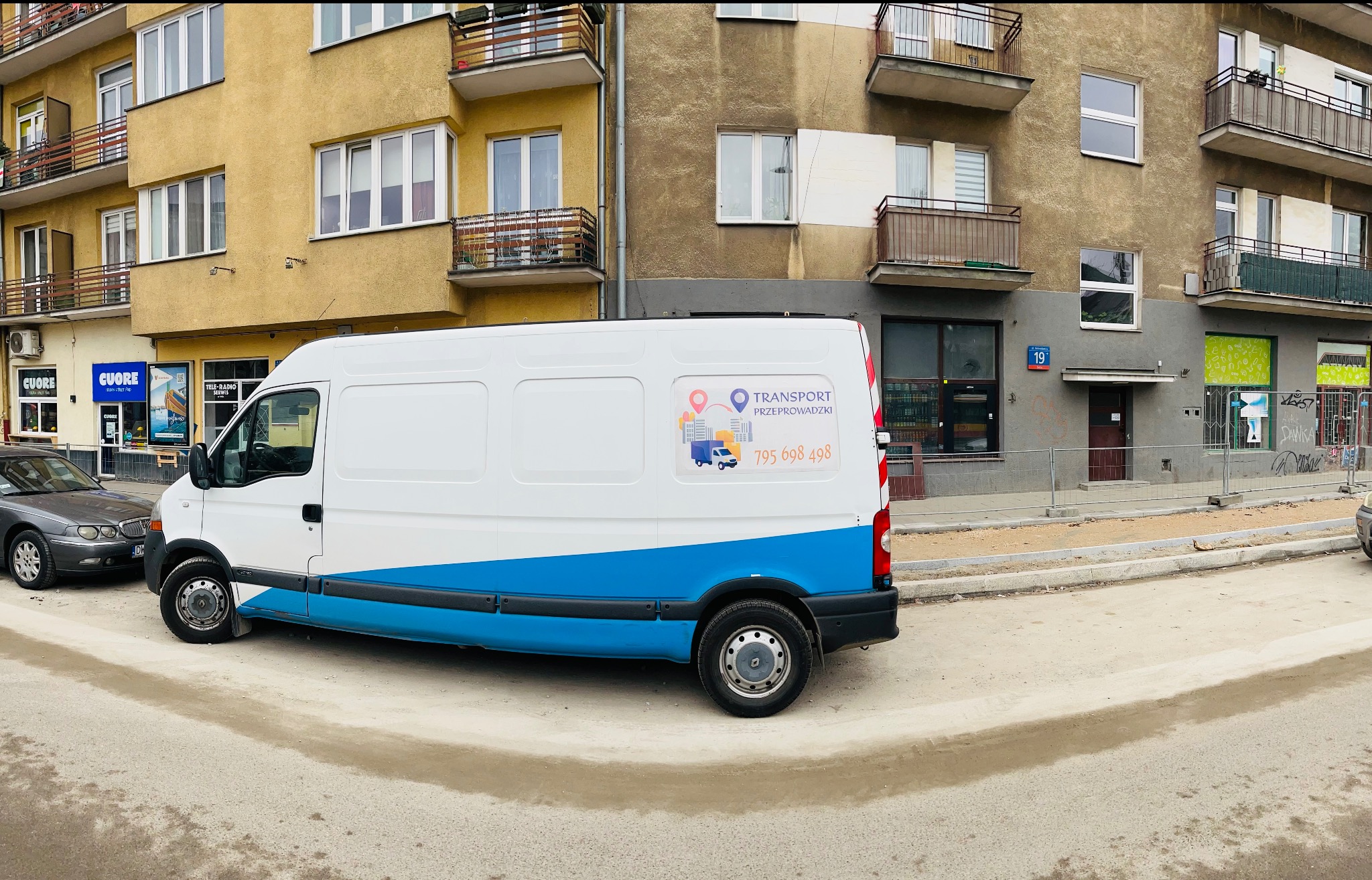 Biała furgonetka z niebieskim pasem i naklejką reklamową firmy transportowej zaparkowana na ulicy przed budynkami mieszkalnymi.