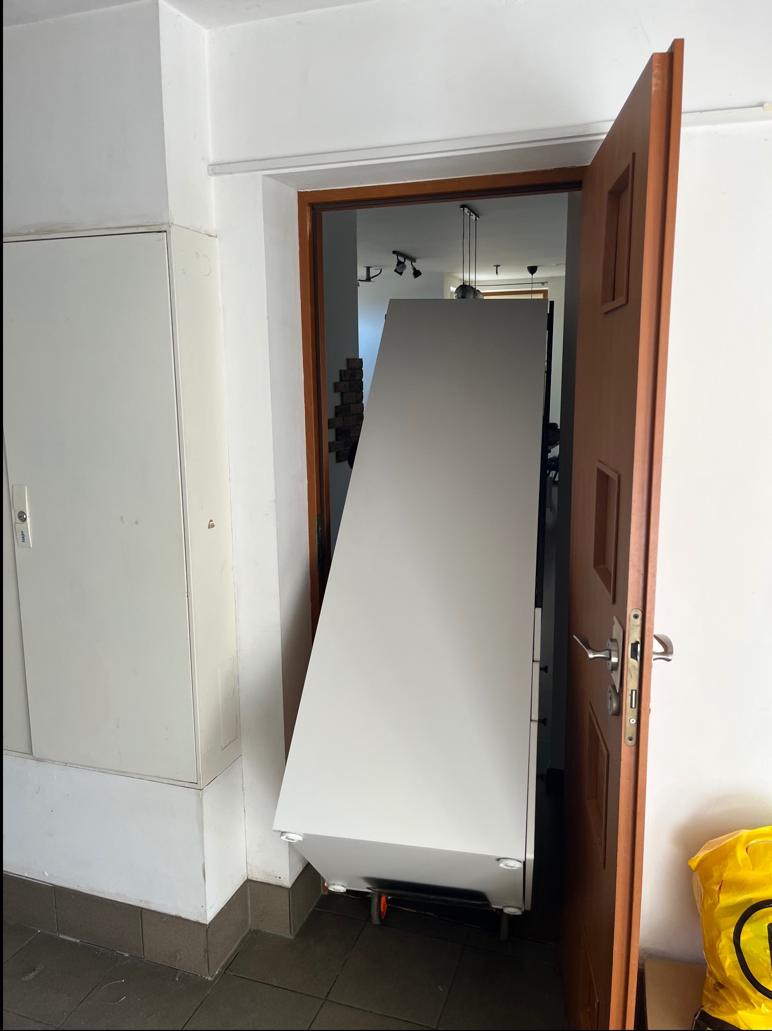 Biała szafa na kółkach, przechylona pod kątem, przechodzi przez futrynę drzwi; widoczny fragment żółtej torby na podłodze.