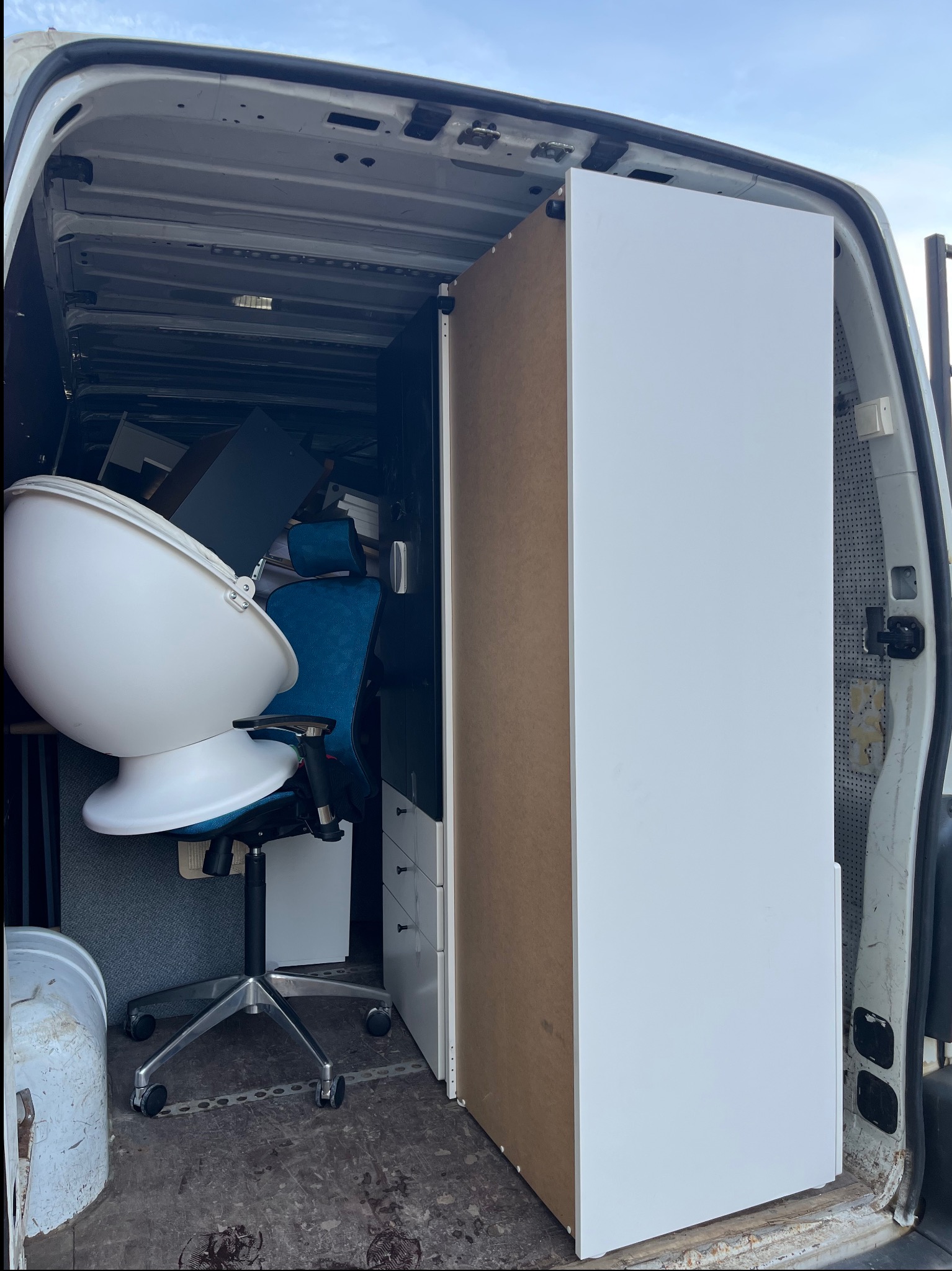 Wnętrze furgonetki wypełnione meblami: krzesło biurowe z białą skorupą, niebieskie krzesło z zagłówkiem, biała szafka z szufladami i inne przedmioty, widoczne ślady użytkowania na podłodze furgonetki.